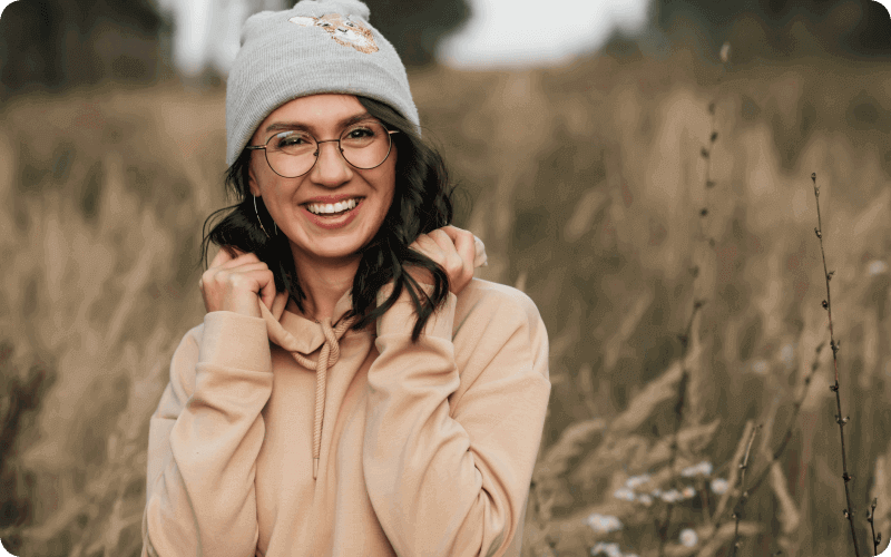 A woman wearing glasses, a hat, and a hoodie in a field. 